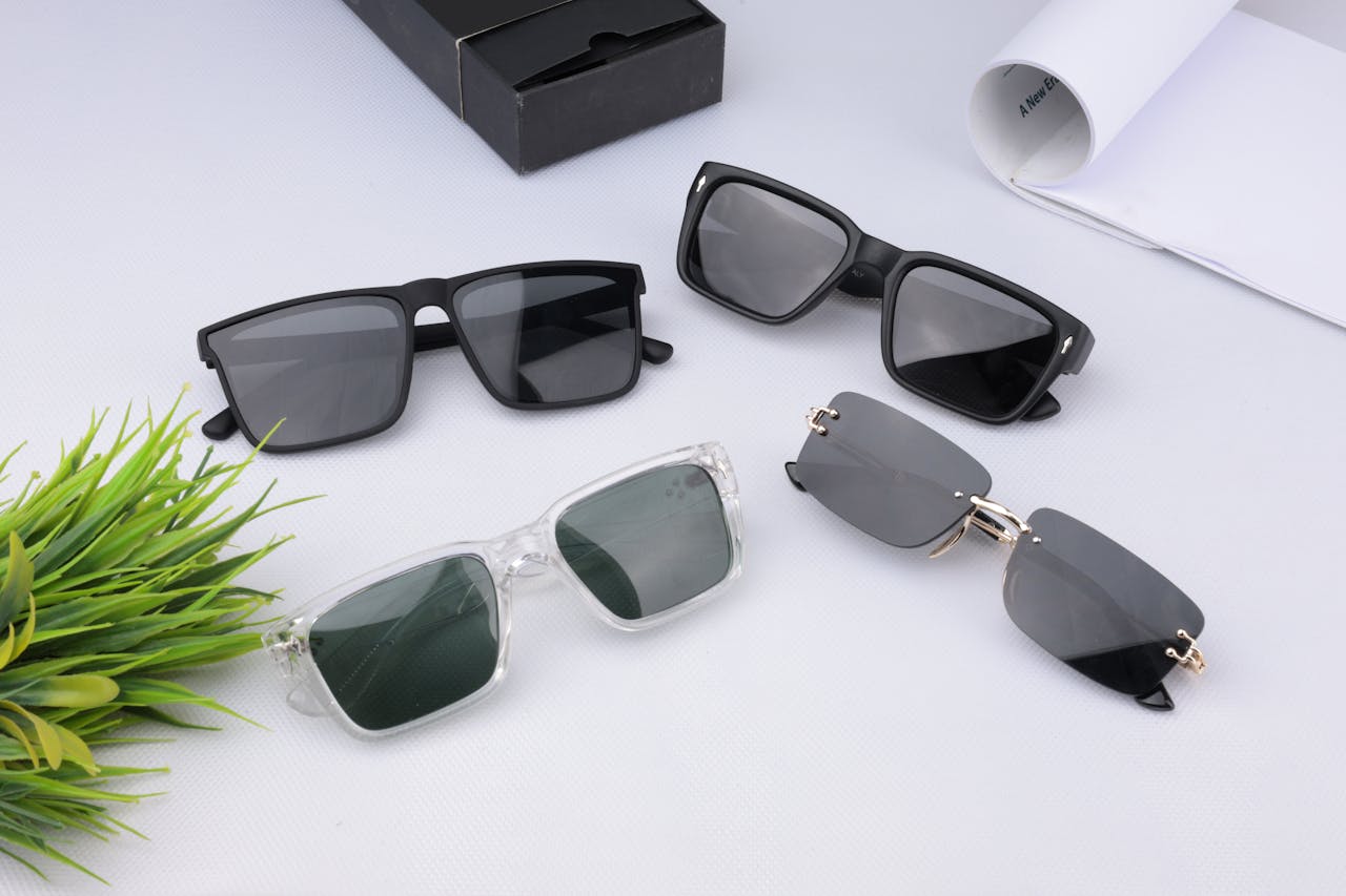 A variety of modern sunglasses laid out on a white surface next to greenery.